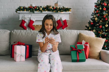 Mädchen im Pyjama mit Handy in der Nähe von Weihnachtsgeschenken und geschmücktem Tannenbaum zu Hause 