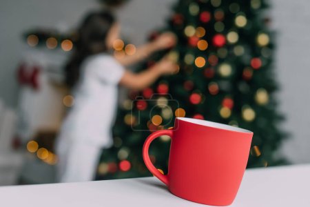 Rote Tasse auf Tisch neben verschwommenem Mädchen und Weihnachtsbaum zu Hause 