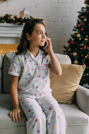 Niño preadolescente en pijama hablando en el teléfono inteligente en el sofá durante la celebración de Navidad en casa 