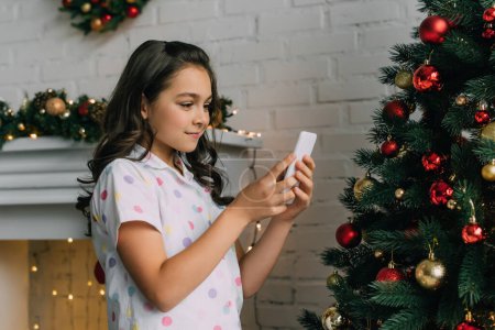Chica preadolescente en pijama usando teléfono inteligente cerca del árbol de Navidad decorado en casa 