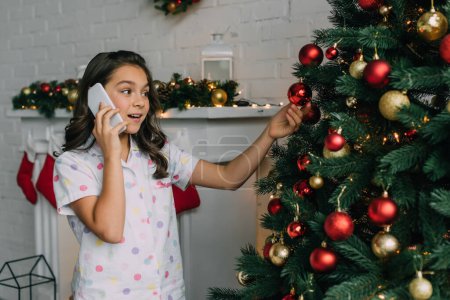 Kid in pajama talking on smartphone and touching ball on Christmas tree 