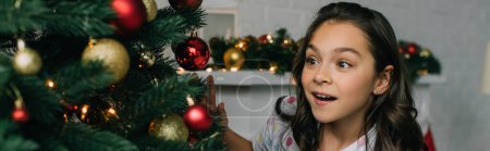 Chica emocionada mirando el árbol de Navidad con adornos en casa, pancarta 