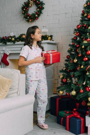 Preteen child in pajama holding present and looking at Christmas tree at home 