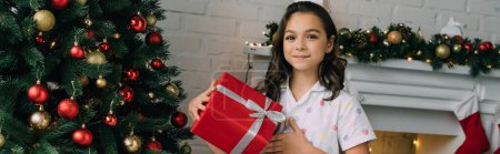 Positives Mädchen im Pyjama hält Geschenk neben Weihnachtsbaum im Wohnzimmer, Banner 