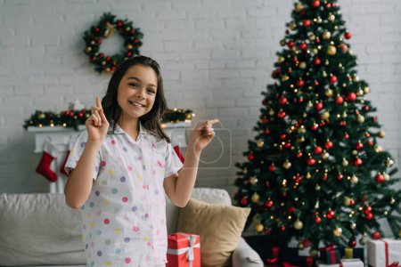 Positive girl in pajama pointing with fingers near blurred Christmas tree in living room 