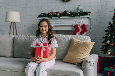 Chica sonriente en pijama punteada sosteniendo regalo mientras se sienta en el sofá durante la celebración de Navidad 