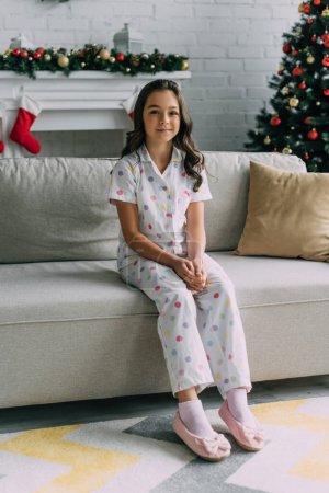 Lächelndes Mädchen im gepunkteten Pyjama auf Couch neben verschwommenem Kamin und Weihnachtsbaum zu Hause 