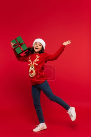 Cheerful girl in Christmas sweater and santa hat holding gift box on red background 