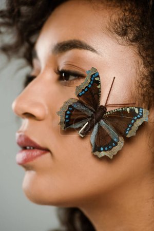 Nahaufnahme von hübschen afrikanisch-amerikanischen Frau mit Schmetterling im Gesicht isoliert auf grau