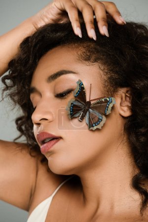 hübsche afrikanisch-amerikanische Frau mit Schmetterling auf Wange isoliert auf grau