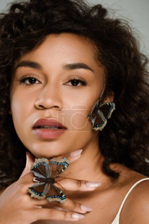 curly african american woman with butterflies on hand and face looking at camera isolated on grey