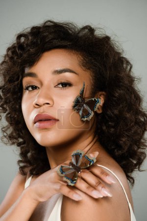african american woman with butterflies on hand and face looking at camera isolated on grey
