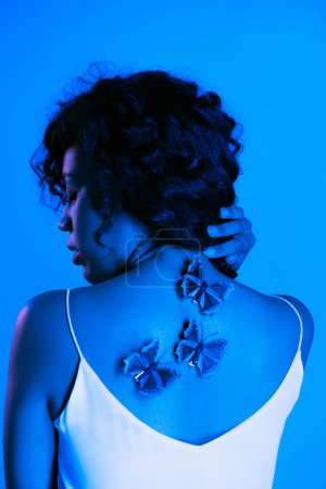 lockige afrikanisch-amerikanische Frau mit Schmetterlingen auf dem Rücken posiert isoliert auf hellblau 