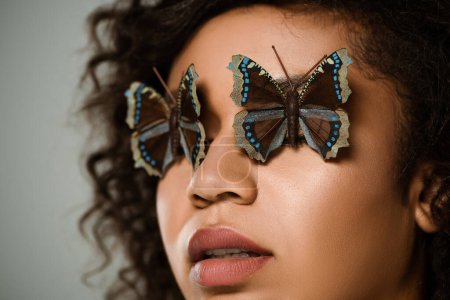 Nahaufnahme einer lockigen afrikanisch-amerikanischen Frau mit Schmetterlingen auf Augen isoliert auf grau