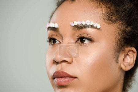 close up of pretty african american woman with flowers on eyebrows isolated on grey