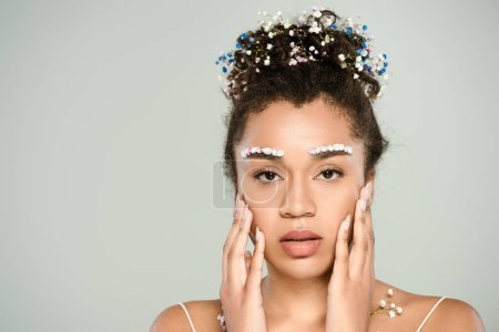 pretty african american woman with flowers on eyebrows and hair touching face isolated on grey