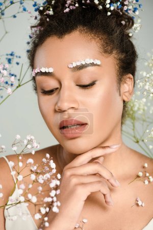 young african american woman with flowers in hair looking down near gypsophila isolated on grey