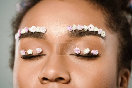 Foto de Vista recortada de mujer afroamericana bonita con flores en las cejas y ojos cerrados aislados en gris - Imagen libre de derechos