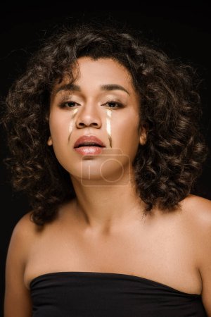 mujer afroamericana con lágrimas de oro en las mejillas mirando a la cámara aislada en negro 