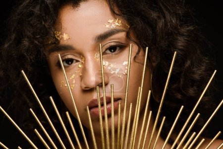curly african american woman with golden paint on cheeks looking at camera through crown isolated on black