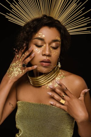 jeune femme afro-américaine avec des accessoires dorés et de la peinture sur le visage posant isolé sur noir