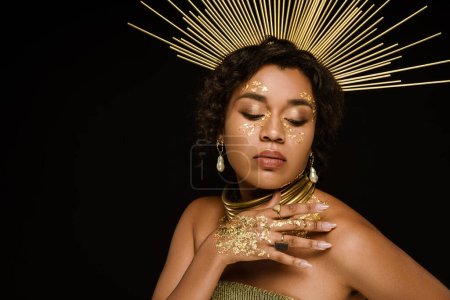 young african american woman with golden accessories and paint on hand posing isolated on black