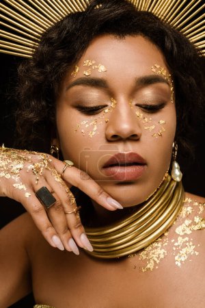 close up of african american woman with golden accessories and paint on face posing isolated on black