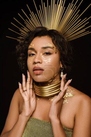 Foto de Joven mujer afroamericana con collar de oro y pintura en la cara mirando a la cámara aislada en negro - Imagen libre de derechos