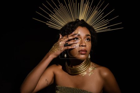 femme afro-américaine tatouée avec couronne dorée et peinture sur le visage regardant isolé sur noir