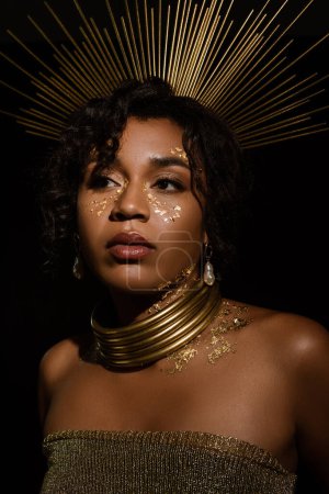mujer afroamericana confiada con corona dorada y pintura en las mejillas mirando hacia otro lado aislado en negro