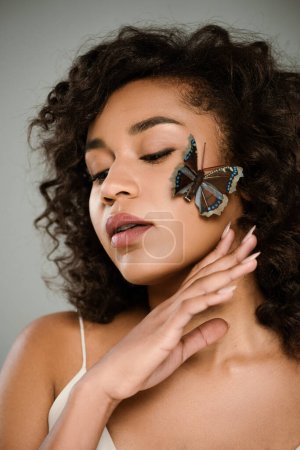 brunette african american woman with butterfly on cheek posing isolated on grey