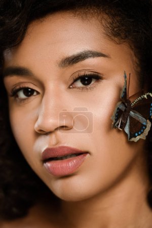 close up of curly african american woman with butterfly on face looking at camera