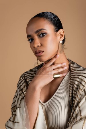 young african american woman in golden jewelry looking at camera isolated on beige