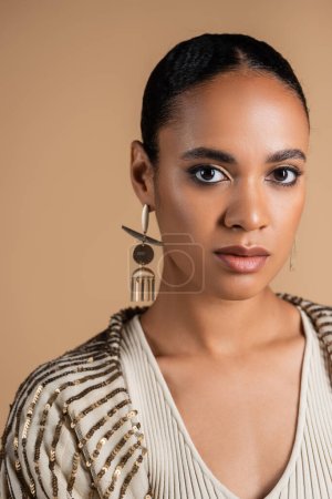 Porträt einer eleganten afrikanisch-amerikanischen Frau mit goldenen Ohrringen, die isoliert auf beige in die Kamera blickt 
