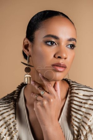 elegant african american woman in golden jewelry touching neck isolated on beige  