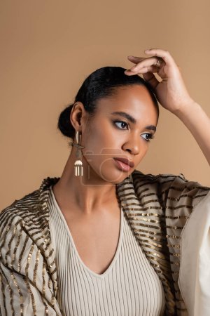 mujer afroamericana con joyas de lujo y chal mirando hacia otro lado aislado en beige 