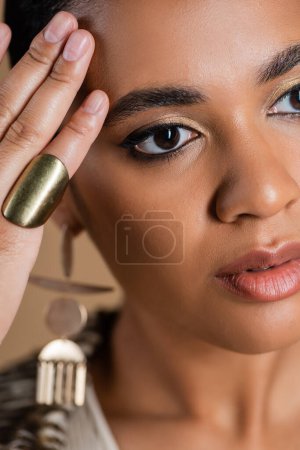 Téléchargez les photos : Gros plan de jeune femme afro-américaine en luxueux bijoux dorés posant isolés sur beige - en image libre de droit