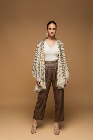 full length of stylish african american woman in golden shawl standing on beige