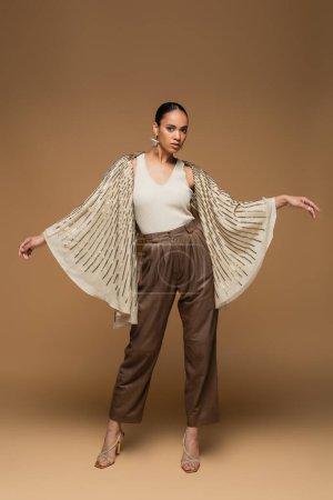 full length of stylish african american woman in golden shawl gesturing on beige