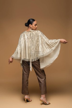full length of stylish african american woman in golden shawl and leather pants standing on beige