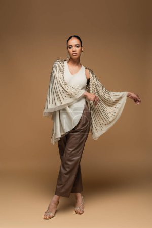 full length of stylish african american woman in golden shawl and leather pants posing on beige
