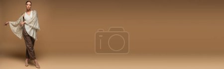 Téléchargez les photos : Longueur totale de femme afro-américaine élégante en châle doré et pantalon en cuir posant sur beige, bannière - en image libre de droit