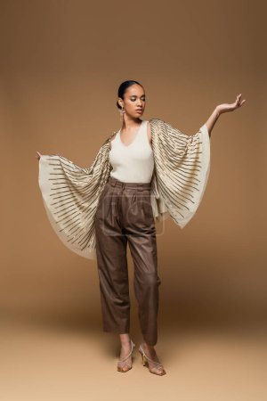 full length of brunette african american woman in golden shawl and leather pants posing on beige 