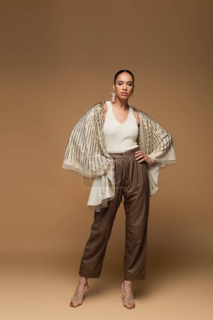 full length of african american woman in golden shawl and leather pants posing with hands on hips on beige