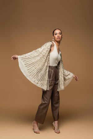 full length of young african american woman in golden jewelry and shawl posing on beige 