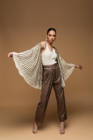 full length of stylish african american woman in golden jewelry and shawl posing on beige 