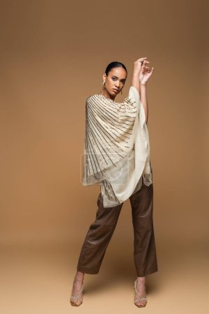 full length of stylish african american model in golden jewelry and shawl posing on beige 