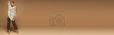 Téléchargez les photos : Pleine longueur de femme brune afro-américaine en bijoux dorés et châle posant sur beige, bannière - en image libre de droit