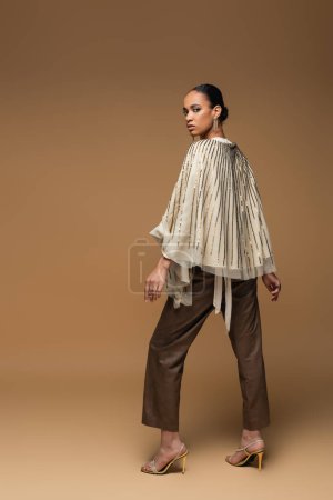 full length of brunette african american woman in luxurious jewelry and golden shawl posing on beige 