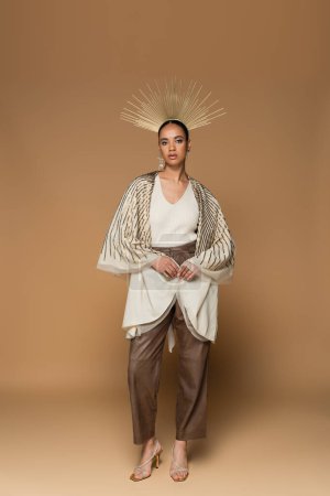 full length of brunette african american woman in shiny shawl and golden crown looking at camera on beige